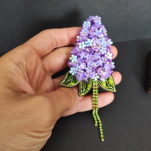 Op de afbeelding: Een paarse en groene kralen broche in de vorm van een bloemcluster. De broche heeft een lange groene kralenketting die aan de onderkant hangt.
