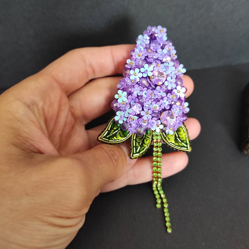 Op de afbeelding: Een paarse en groene kralen broche in de vorm van een bloemcluster. De broche heeft een lange groene kralenketting die aan de onderkant hangt.