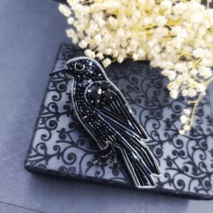 May include: A black beaded crow brooch with a shiny black finish. The brooch is detailed with black beads and a black crystal eye.