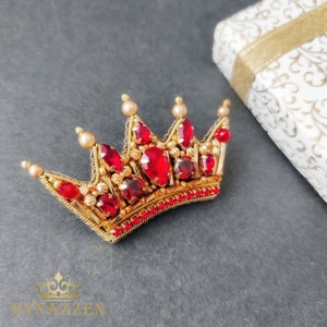 Op de afbeelding: Een gouden kroon met rode juwelen en parel accenten. De kroon is een handgemaakte broche.