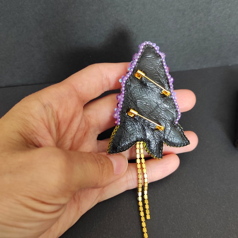Op de afbeelding: Een broche in de vorm van een kerstboom met paarse en gele kralen, een zwarte vilt achterkant en gouden kettingaccenten. De broche heeft twee veiligheidsspelden aan de achterkant om hem aan kleding te bevestigen.