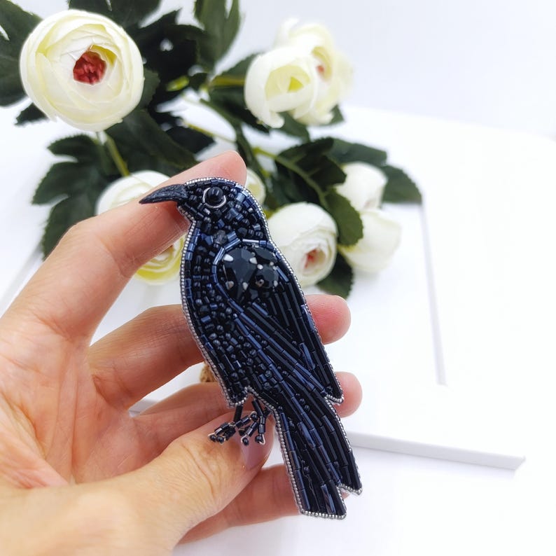 May include: A black beaded crow brooch with a dark blue iridescent finish. The brooch is detailed with black beads and has a silver pin on the back.