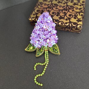 Op de afbeelding: Een paarse en groene kralen broche in de vorm van een bloemcluster. De broche is gemaakt met kleine paarse kralen en groene kralen, en heeft een lange groene kralenketting die aan de onderkant hangt.