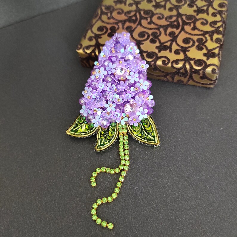 Op de afbeelding: Een paarse en groene kralen broche in de vorm van een bloemcluster. De broche is gemaakt met kleine paarse kralen en groene kralen, en heeft een lange groene kralenketting die aan de onderkant hangt.