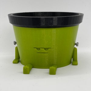 May include: A green 3D printed planter with a black rim and small legs. The planter has a grumpy face with two eyebrows.