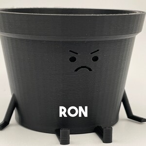 Puede incluir: Una maceta de plástico negra con una cara de enfadado y la palabra "RON" impresa en la parte delantera. La maceta tiene cuatro pequeñas patas.
