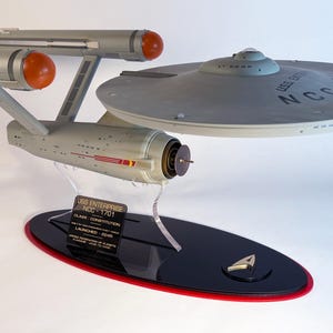 May include: A model of the USS Enterprise NCC-1701 starship, a science fiction icon. The model is gray with orange engine nacelles and red accents. It sits on a black base with a red trim and a Starfleet insignia.