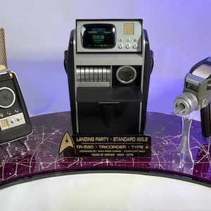 Może przedstawiać: Zestaw rekwizytów Star Trek zawierający czarno-złoty komunikator, czarno-srebrny tricorder TR-530 oraz srebrno-czarny pistolet phaser. Przedmioty umieszczono na zakrzywionej fioletowej podstawie z wzorami gwiazdozbiorów i trzema czarnymi tabliczkami.