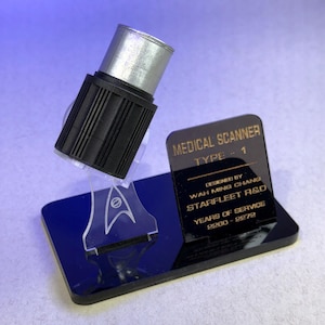 Puede incluir: Un escáner médico negro y plateado, Tipo 1, con una base con el emblema de Starfleet. El accesorio incluye el texto "MEDICAL SCANNER TYPE - 1" y "YEARS OF SERVICE 2260-2272". Diseñado por Wah Ming Chang, Starfleet R&D.