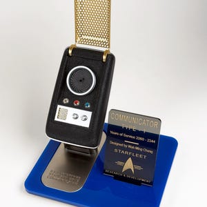 May include: A black and gold Star Trek Type-1 Communicator replica, with a blue base and a silver stand. The communicator has a gold perforated top and a circular speaker. A blue plaque displays text about the communicator.