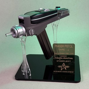 Peut inclure: Réplique d'un pistolet Phaser Type-2 noir et gris, présentée sur un socle noir avec des supports en acrylique transparent. L'accessoire présente des accents argentés et le logo Starfleet Research & Development. Le texte sur le socle indique "Phaser Pistol Type-2".