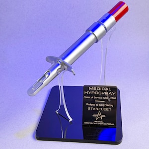 Puede incluir: Una réplica de utilería plateada de Medical Hypospray con punta roja, exhibida sobre un soporte transparente y una base negra. La base presenta una placa con el texto "MEDICAL HYPOSPRAY" y "STARFLEET".