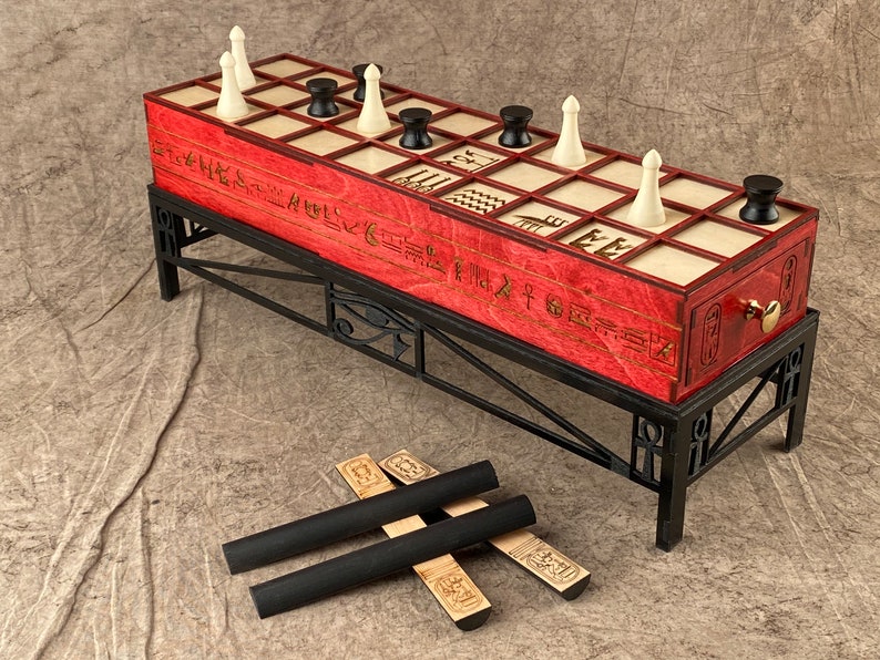 Puede incluir: Un tablero de juego de madera roja con piezas blancas y negras, jerogl&iacute;ficos y un soporte de metal negro. El tablero de juego est&aacute; sobre una superficie gris con tres palos negros y de madera.