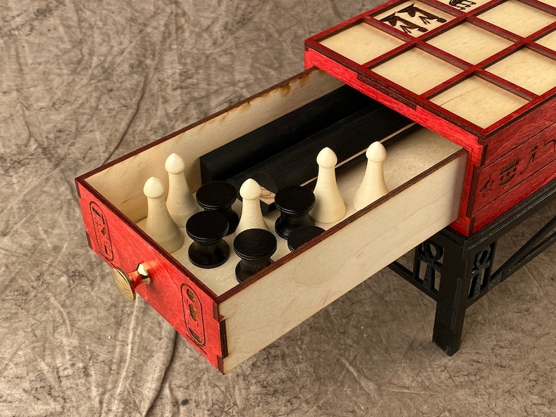 Puede incluir: Un tablero de juego de madera con un acabado rojo y negro. El tablero tiene un caj&oacute;n que se abre para revelar piezas de juego blancas y negras y dos palos negros. El tablero est&aacute; sobre un soporte negro con un dise&ntilde;o de jerogl&iacute;ficos egipcios.