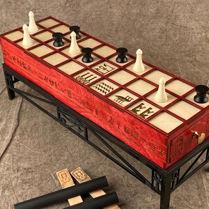 Puede incluir: Un tablero de juego de madera roja con piezas blancas y negras sobre un soporte de metal negro. El tablero tiene jerogl&iacute;ficos y s&iacute;mbolos tallados. El juego se llama "Senet" y es un antiguo juego de mesa egipcio.