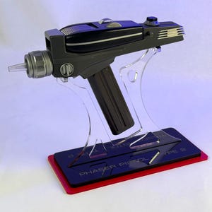 May include: A black and gray phaser pistol prop with a silver emitter tip, mounted on a clear acrylic stand. The base features a black plate with the text PHASER PISTOL - TYPE 2 printed in white, resting on a red rectangular platform.