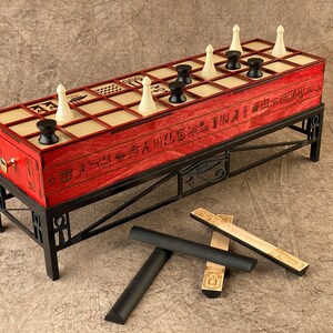 Puede incluir: Un tablero de juego de madera roja con piezas blancas y negras, colocado sobre un soporte de metal negro. El tablero tiene jerogl&iacute;ficos y la inscripci&oacute;n "Senet Game". Hay cuatro palos de madera con jerogl&iacute;ficos en el suelo.