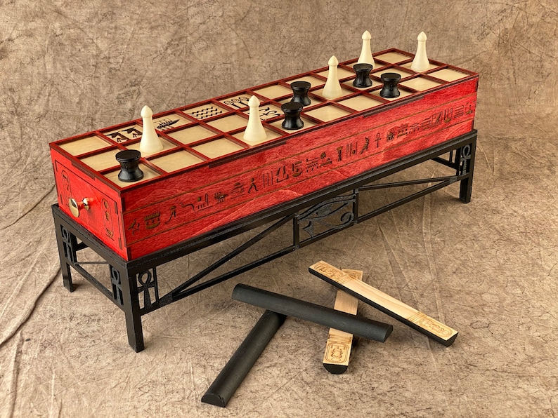 Puede incluir: Un tablero de juego de madera roja con piezas blancas y negras, colocado sobre un soporte de metal negro. El tablero tiene jerogl&iacute;ficos y la inscripci&oacute;n "Senet Game". Hay cuatro palos de madera con jerogl&iacute;ficos en el suelo.
