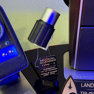 May include: A black and silver medical scanner prop with the text "MEDICAL SCANNER TYPE - 1" and "DESIGNED BY W. M. CHANG". The prop also includes the text "YEARS OF SERVICE 2200-2279". Other props are visible in the background.