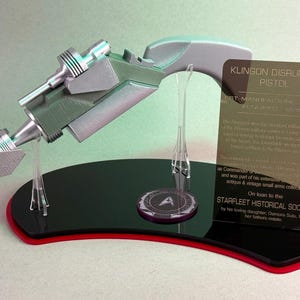 Może przedstawiać: Srebrna i szara replika pistoletu Klingon Disruptor, eksponowana na czarnej i czerwonej podstawie z emblematem Starfleet Historical Society. Zawiera przezroczystą akrylową kartę informacyjną. Kolekcjonerski przedmiot science fiction.
