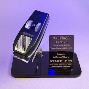 Peut inclure: Réplique d'un Hand Phaser Type-1 noir avec des détails argentés, présenté sur un socle noir. Le socle porte une plaque avec le texte "Hand Phaser Type-1 Standard Issue". Conçu par Jefferies/Chang, cet objet est de collection.