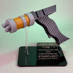 May include: A gray and orange laser pistol, Type 1, on a clear stand with a black base. The pistol has a ribbed handle and a cylindrical barrel. The base has text that reads "LASER PISTOL TYPE - 1" and "STARFLEET RESEARCH & DEVELOPMENT."