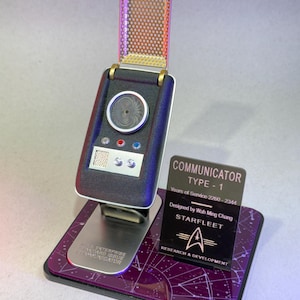 May include: A black and silver Star Trek Type-1 Communicator prop replica. The prop has a flip-up top with a gold mesh grill. A stand with a purple base and text that reads "COMMUNICATOR TYPE-1" is included.