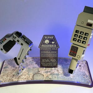 May include: A silver stun gun and a beige commlock device displayed on a stand with a lunar map base. A central plaque reads ALPHA MOONBASE STANDARD ISSUE COMMLOCK and STUN GUN LASER WEAPON MODEL 2, with additional technical text detailing the equipment.