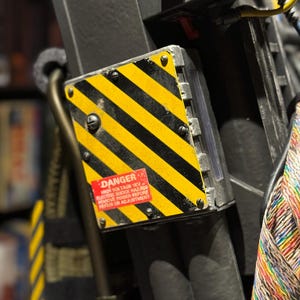 May include: Close-up of a black and yellow striped panel with the word "DANGER" in red. The panel is part of a larger, dark-colored prop, possibly a movie or cosplay item. The text on the panel warns of high voltage and electric shock hazards.
