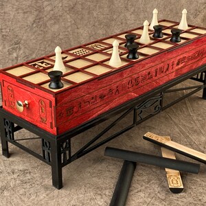 Puede incluir: Un tablero de juego de madera roja con piezas blancas y negras, colocado sobre un soporte de metal negro con un dise&ntilde;o egipcio. El tablero tiene jerogl&iacute;ficos en el lateral y el soporte tiene un dise&ntilde;o de Ojo de Horus. Tambi&eacute;n hay cuatro palos de madera con jerogl&iacute;ficos en ellos.