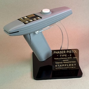 Puede incluir: Réplica gris de una pistola Phaser Tipo-2, montada sobre una base negra. El accesorio presenta un botón blanco, una serie de paneles rectangulares y el texto "PHASER PISTOL TYPE - 2" y "STARFLEET RESEARCH & DEVELOPMENT".