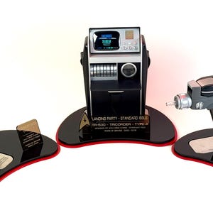 May include: Three Star Trek prop replicas: a communicator, a tricorder, and a phaser pistol. Each prop is displayed on a black base with a red trim. The communicator is black and gold, the tricorder is black and silver, and the phaser is gray and black.