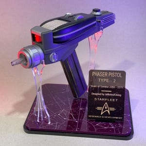 May include: A black and gray Phaser Pistol Type-2 replica, displayed on a purple base with a constellation design. The prop features a red light and the Starfleet logo. The text on the base reads "PHASER PISTOL TYPE-- 2".