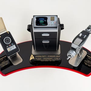 Puede incluir: Un conjunto de réplicas de accesorios de Star Trek que incluye un comunicador, un tricorder y una pistola de fase. El conjunto se muestra sobre una base negra y roja. El tricorder muestra "16:52:04".