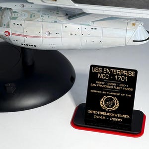 Puede incluir: Maqueta de la nave espacial USS Enterprise NCC-1701, con un soporte de exhibición negro y rojo. El soporte muestra el nombre, el registro y los detalles de servicio de la nave. El modelo es blanco con detalles en negro y rojo.
