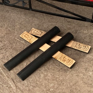 Puede incluir: Dos varillas de madera negra con extremos redondeados se cruzan sobre dos tiras de madera con dise&ntilde;os jerogl&iacute;ficos. Las tiras est&aacute;n grabadas con el texto "ECO2".