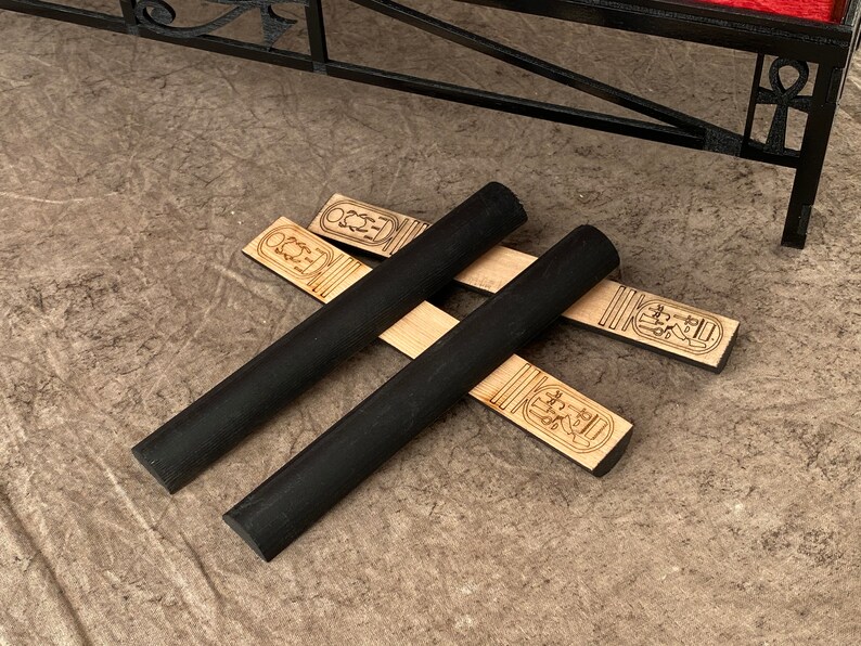 Puede incluir: Dos varillas de madera negra con extremos redondeados se cruzan sobre dos tiras de madera con dise&ntilde;os jerogl&iacute;ficos. Las tiras est&aacute;n grabadas con el texto "ECO2".