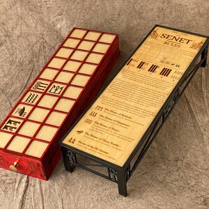 Puede incluir: Un tablero de juego de Senet de madera con ribete rojo y un soporte de metal negro con reglas impresas en la superficie. El tablero de juego tiene un caj&oacute;n con una manija de lat&oacute;n. Las reglas incluyen el texto "The Game of Senet Rules".