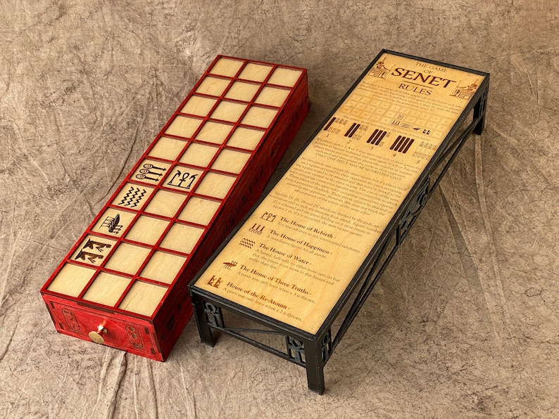 Puede incluir: Un tablero de juego de Senet de madera con ribete rojo y un soporte de metal negro con reglas impresas en la superficie. El tablero de juego tiene un caj&oacute;n con una manija de lat&oacute;n. Las reglas incluyen el texto "The Game of Senet Rules".