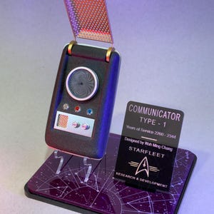 May include: A black and silver Star Trek Type-1 communicator replica with an open top, mounted on a purple base. The base has a constellation pattern and a Starfleet logo. The communicator has a speaker, buttons, and a small screen. A sign reads "Communicator Type-1".