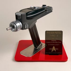 May include: A black and silver Phaser Pistol Type-2 replica, displayed on a red base with a silver stand. The prop features a detailed design and is accompanied by a plaque with text that reads "PHASER PISTOL TYPE-2".