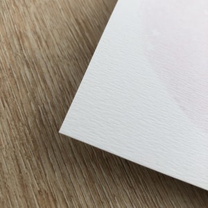May include: A close-up of a white textured card stock paper. The paper has a subtle grain pattern and is slightly off-white in color.