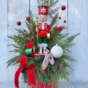 May include: A decorative nutcracker figure wearing a red and green uniform with a snow globe on its head, placed in a container with evergreen branches, red glittery spheres, a white ornament, a red velvet ribbon, and pink ballet slippers.