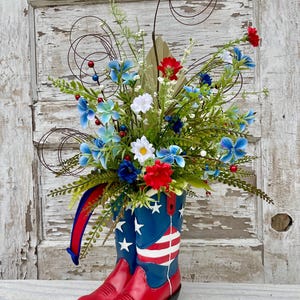 May include: A patriotic floral arrangement in a pair of red cowboy boots. The boots are decorated with the American flag. The arrangement includes blue, white, and red flowers, greenery, and decorative swirls, perfect for a Fourth of July celebration.