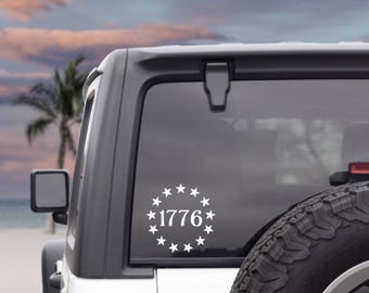 May include: A white vinyl decal applied to a vehicle window featuring the number 1776 centered within a circular arrangement of thirteen five-pointed stars. The design is monochromatic and displays clean, bold lines against the dark glass surface.