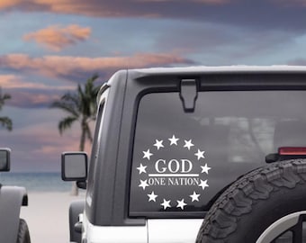 May include: White decal on a vehicle's rear window. The decal features a circle of stars surrounding the words "GOD" and "ONE NATION" in a bold, clean font. The background shows a sunset over the ocean.