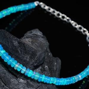 Puede incluir: Un delicado brazalete hecho de cuentas de ópalo azul claro con un cierre plateado. El brazalete se muestra sobre una roca gris oscura.