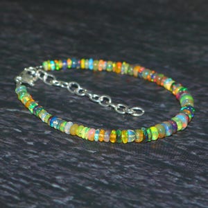 Op de afbeelding: Een kleurrijke armband met opaal edelstenen. De armband heeft een zilveren ketting en bestaat uit kleine, ronde, veelkleurige kralen. De kralen tonen tinten groen, oranje, geel, blauw en paars. De armband is ongeveer 18 cm lang.