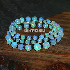 May include: A three-strand bracelet featuring iridescent opal beads in shades of blue, green, and white. The beads are strung on black cord and secured with a silver clasp. The text "ETHIOPIANOPALHUB" is visible.