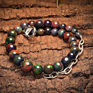Op de afbeelding: Een armband van zwarte opaal kralen met een zilveren sluiting en ketting. De armband ligt op een bruin houten oppervlak.
